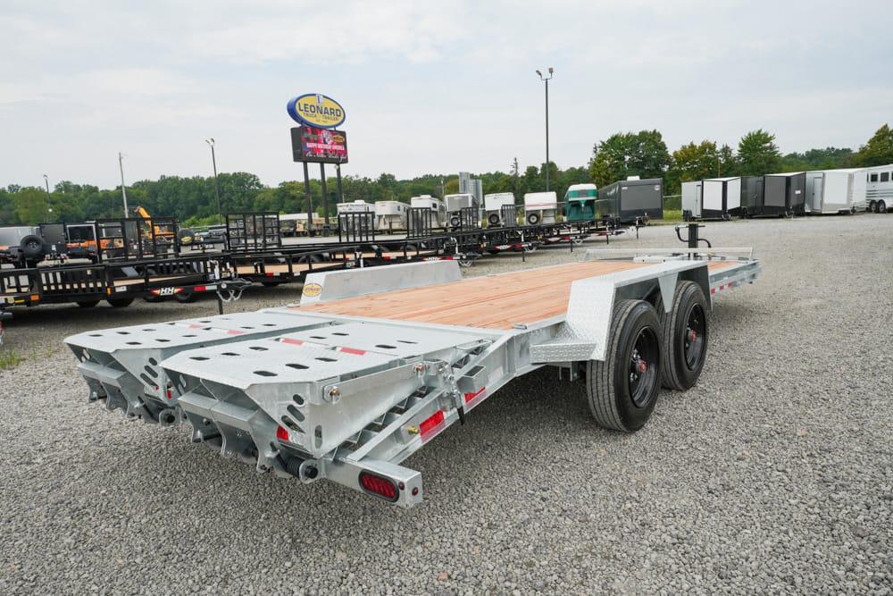 Dual-axle metal flatbed trailer with a wooden deck on a gravel lot, surrounded by other trailers and equipment in the background.