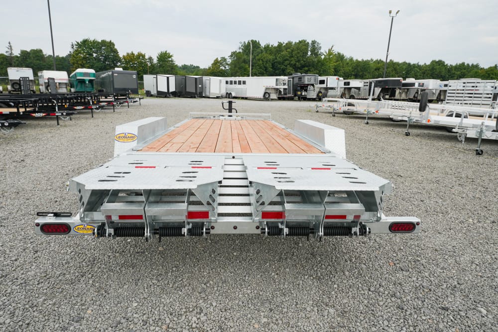 Wide view of a metal car hauler trailer with a wooden deck parked in a gravel lot among other trailers; rear ramps folded up and taillights visible, ready to haul vehicles.