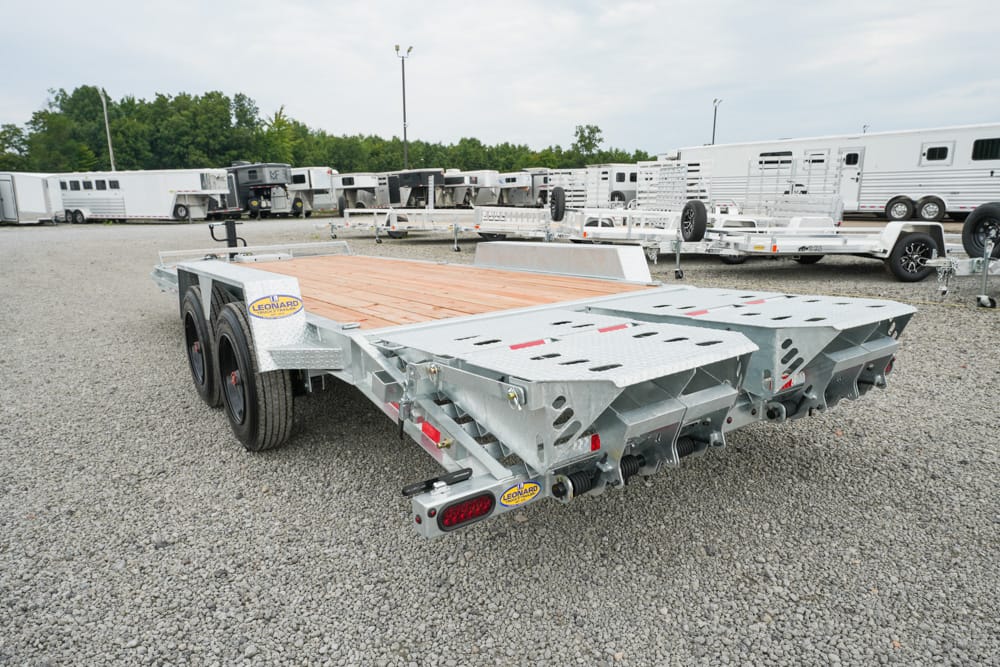 Heavy aluminum flatbed trailer with wooden deck parked in a gravel lot among other trailers; dual rear axles and hook-up area visible.