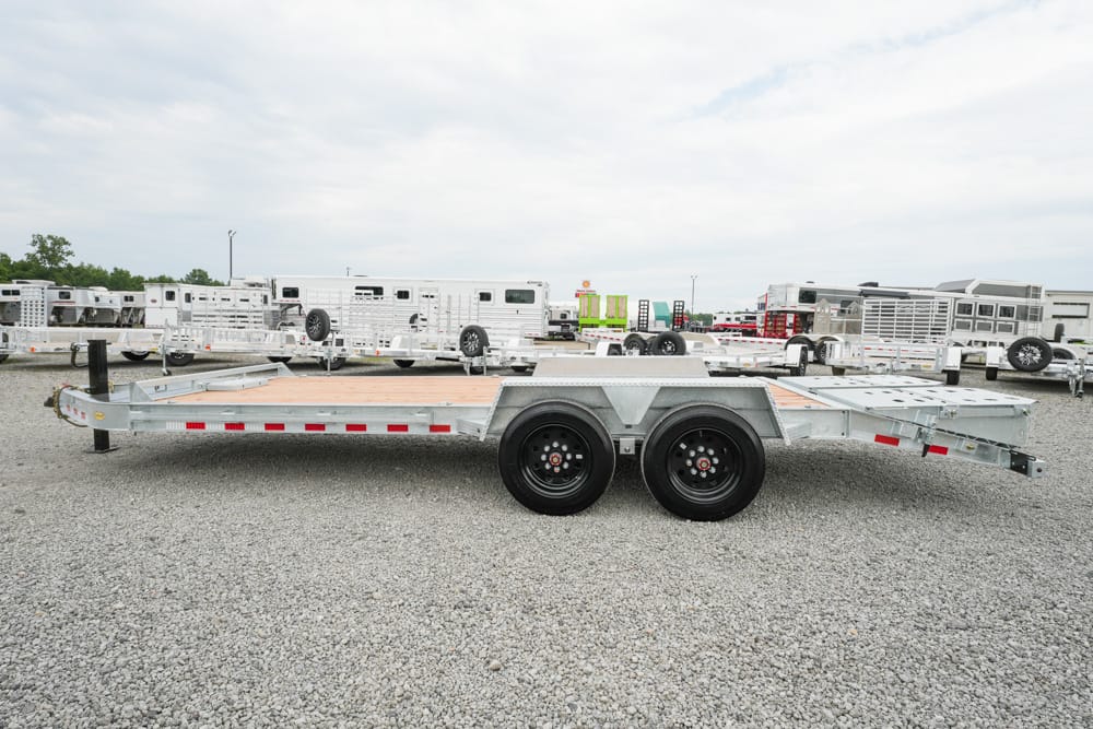 Dual-axle flatbed trailer with a wooden deck on a gravel lot, surrounded by other trailers and equipment.