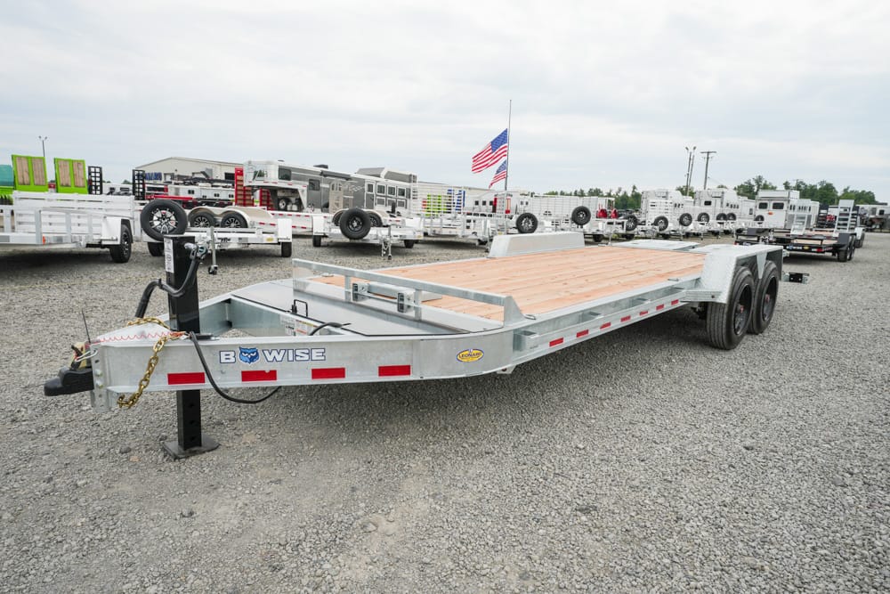 Flatbed trailer with a wooden deck, dual rear axles, and a hitch, parked on a gravel lot with other trailers in the background and an American flag nearby.