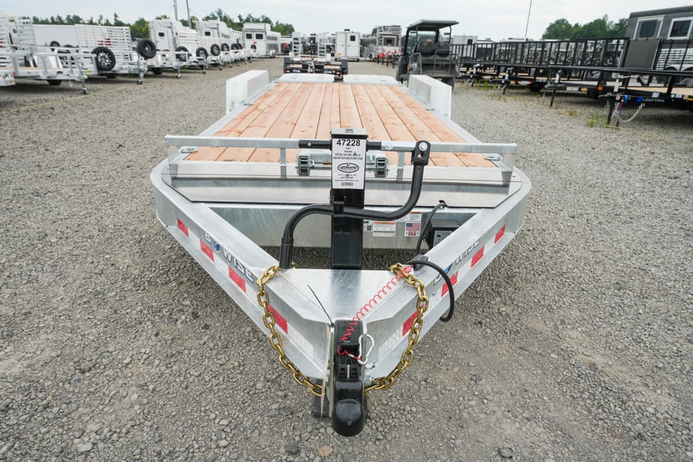 Front view of a silver utility trailer with a wood deck and safety chains, parked on a gravel lot with other trailers in the background.