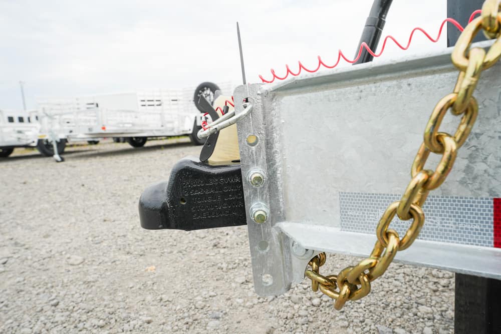 Close-up of a metal trailer hitch with a chain and a red safety cord on a gravel lot, with other trailers in the background.