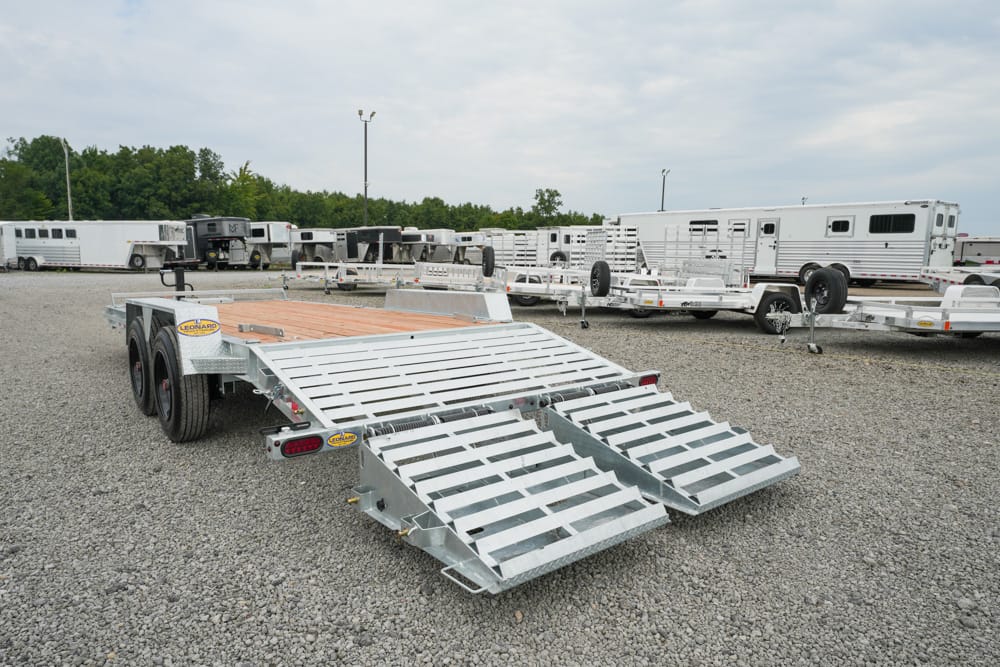 Aluminum car hauler trailer with loading ramps extended in a gravel lot, other trailers in the background.