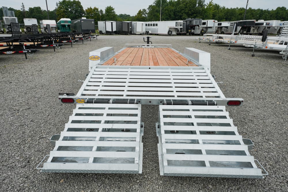 Dual-ramp aluminum flatbed trailer in a gravel lot, ready for loading with a wooden deck surface at the rear area of a trailer yard.