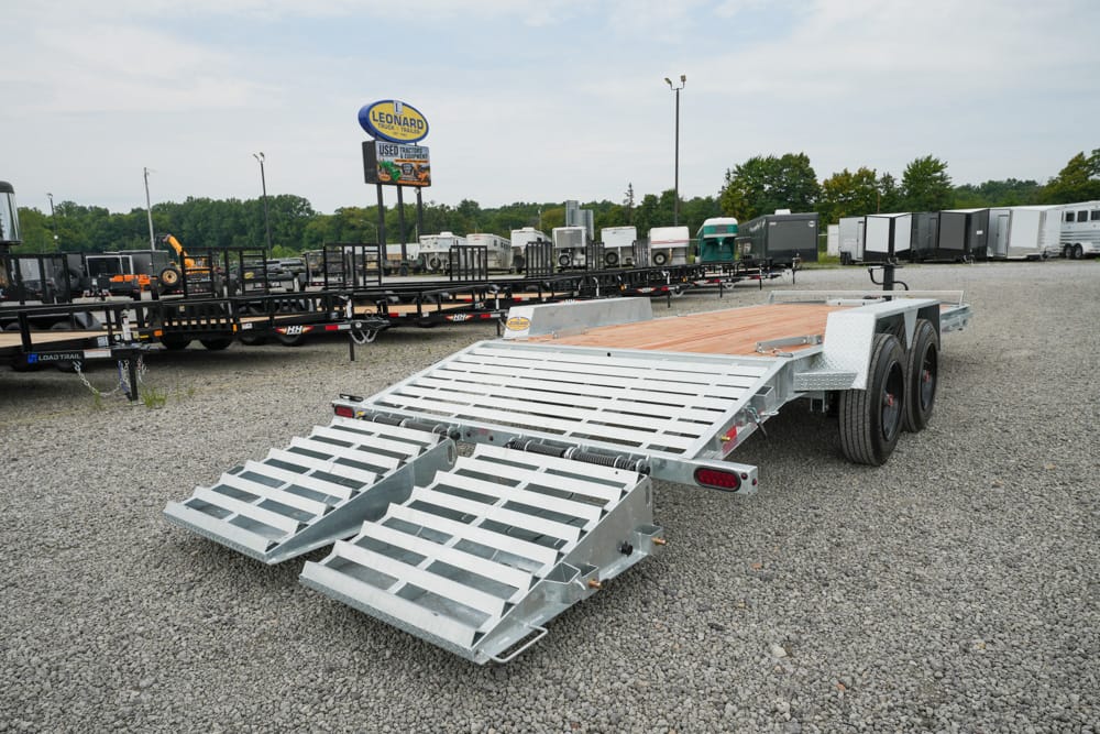 Dual-axle tilt-bed trailer with two large folding ramps extended, parked in a gravel lot with other trailers nearby.