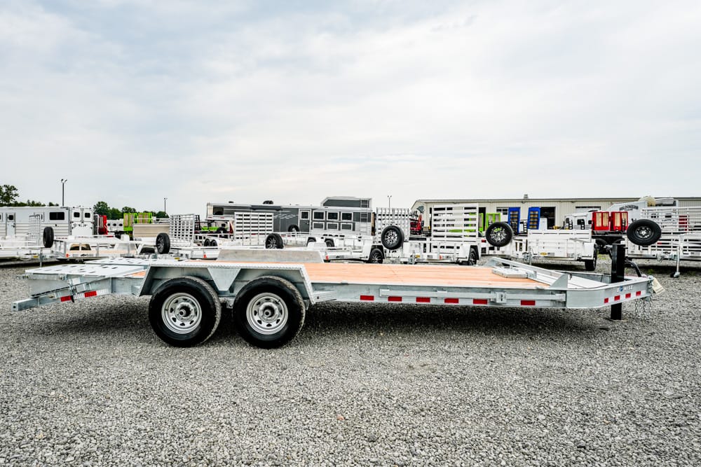 A long flatbed trailer with dual rear axles sits on a gravel lot among other trailers in the background.