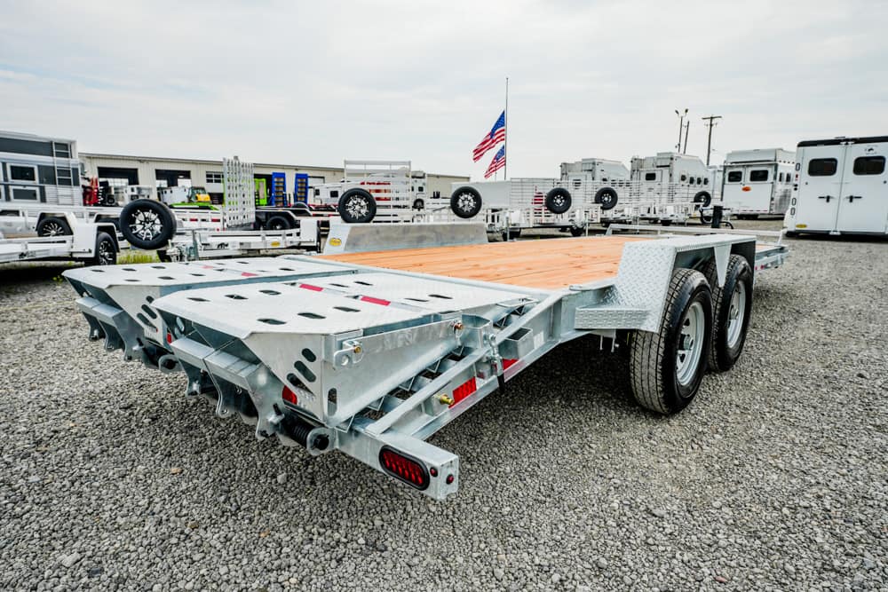 Dual-axle aluminum car trailer with a wooden deck parked on a gravel lot, surrounded by other trailers and equipment. A waving American flag is visible in the background.