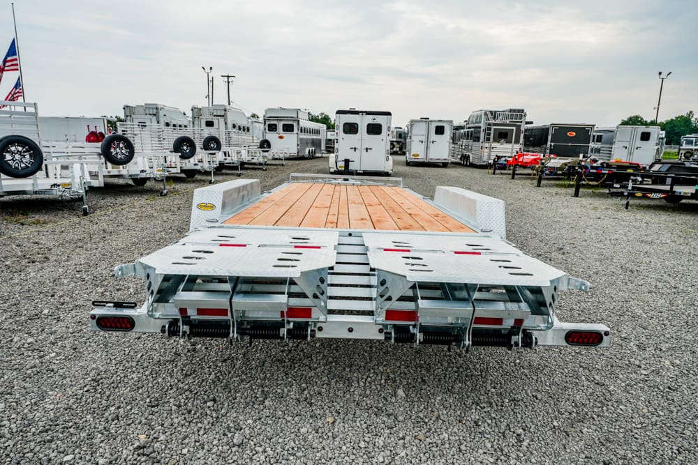 Flatbed trailer with a wooden deck and metal ramps in a gravel lot, surrounded by horse trailers in the background.