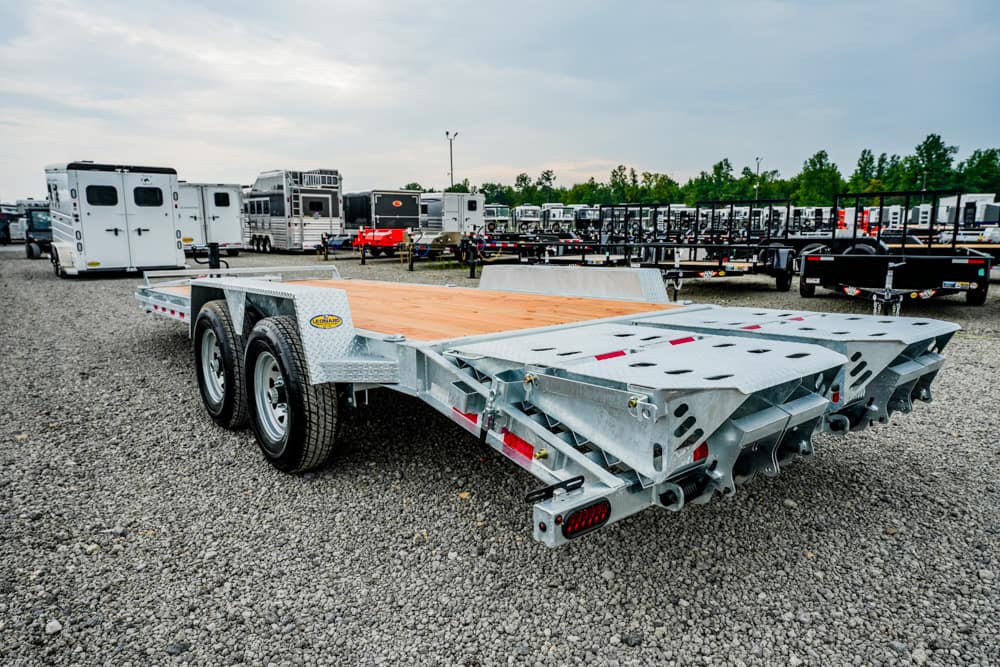 Silver dual-axle flatbed trailer on a gravel lot with other trailers in the background, wooden deck and metal ramps visible on the rear.