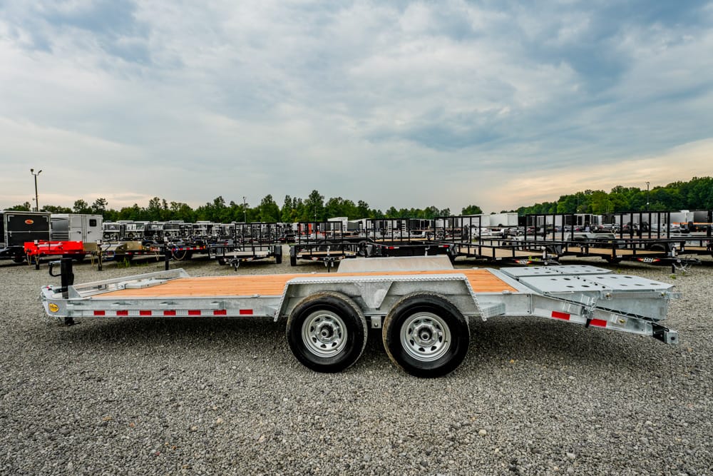 Side view of a silver dual-axle flatbed trailer with a wooden deck in a gravel lot among many trailers