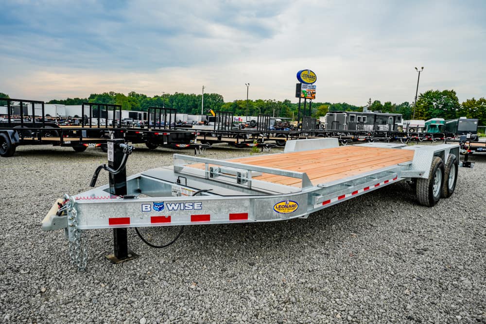Silver two-axle flatbed trailer with a wooden deck, parked in a gravel lot among other trailers