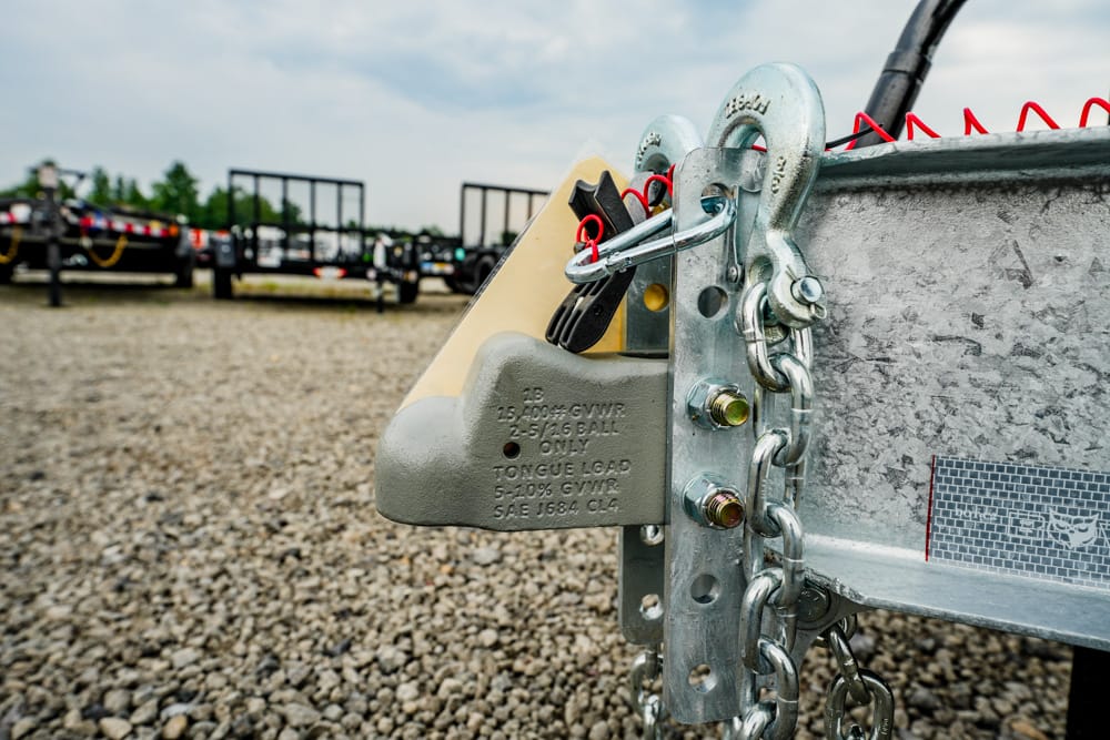 Close-up of a trailer hitch lock and chain securing the tongue of a metal trailer on a gravel lot, with other trailers in the background.