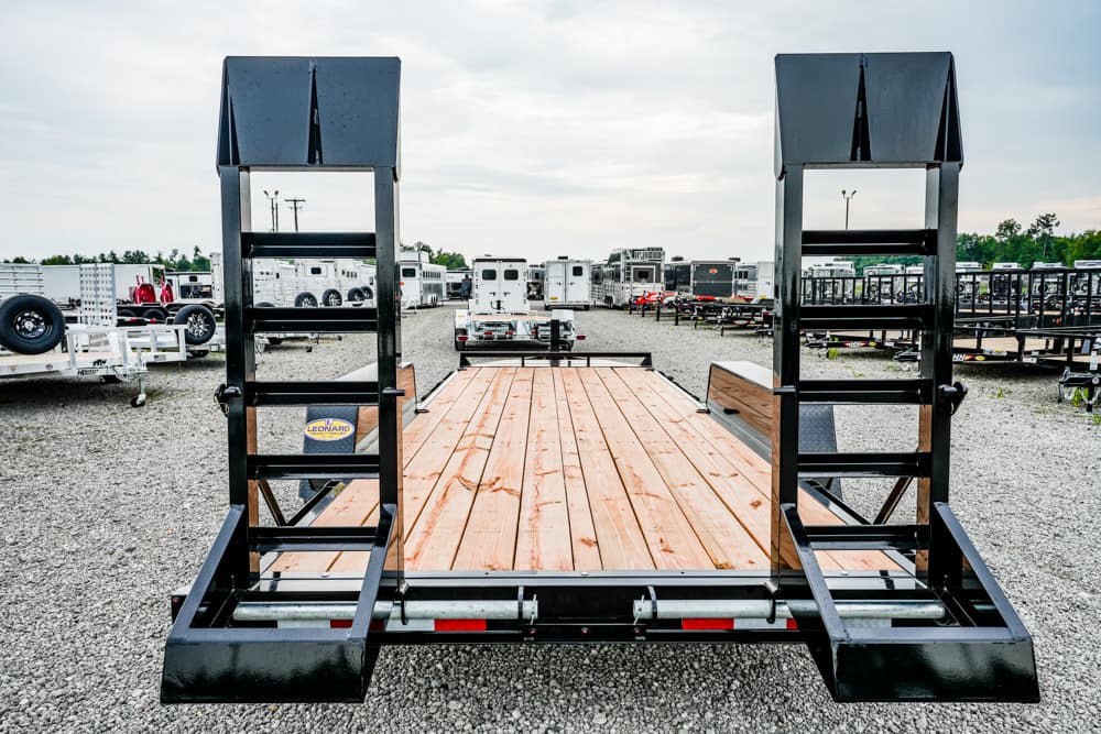 Wide view of a black flatbed trailer with a wooden deck and two tall metal ramps at the rear, parked in a gravel trailer yard.
