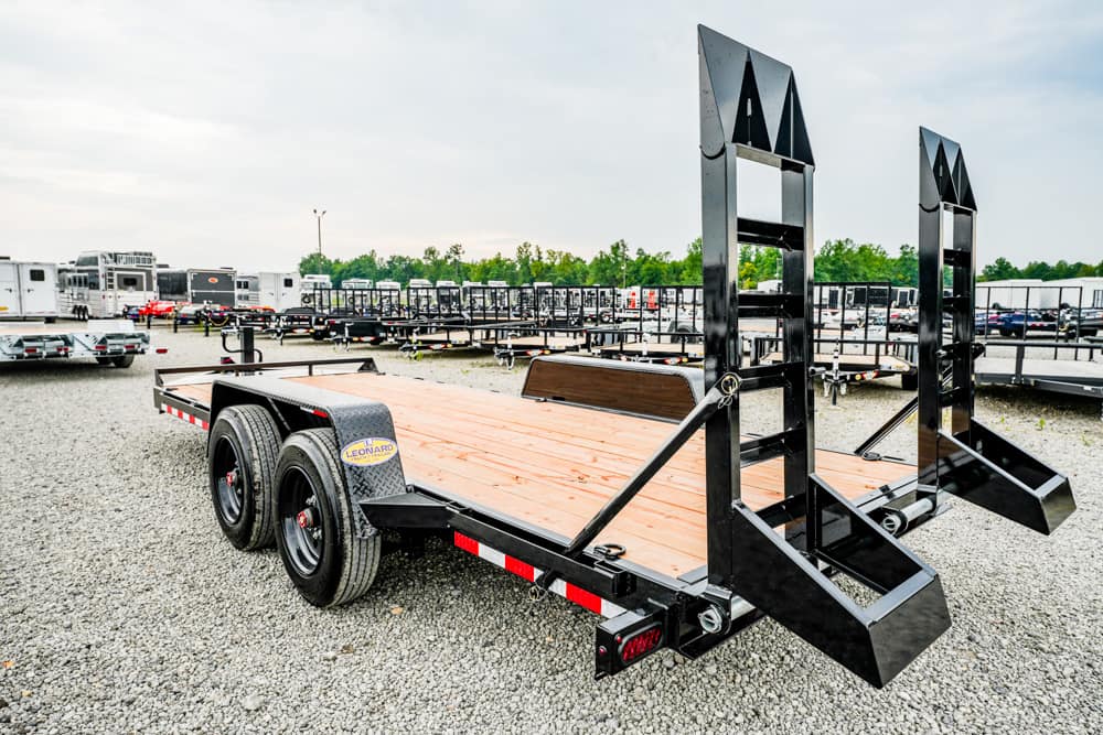 Black flatbed trailer with a wooden deck and two fold-down ramps in a gravel trailer lot.