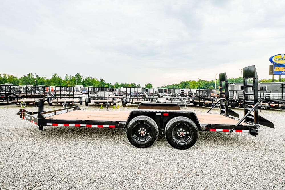Flatbed trailer with dual axles parked on a gravel lot among other trailers in a dealership yard.