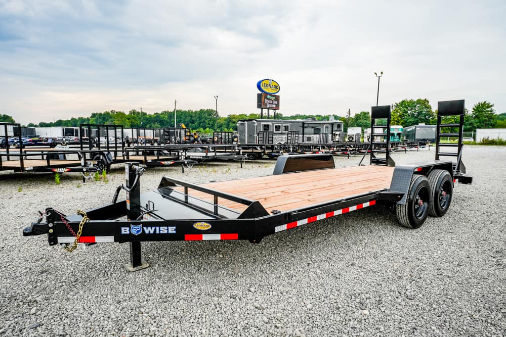 Black B-WISE flatbed trailer with a wooden deck and dual-axle setup on a gravel lot at a Leonard trailer dealership, with loading ramps at the rear.