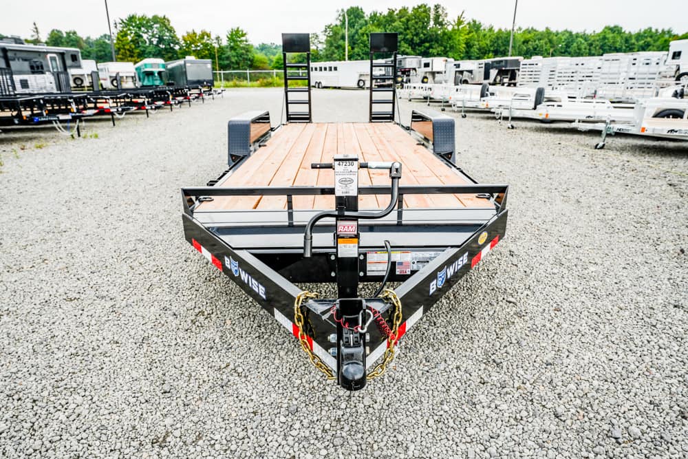 Front view of a black steel flatbed trailer with a wooden deck, parked on a gravel lot with other trailers nearby.
