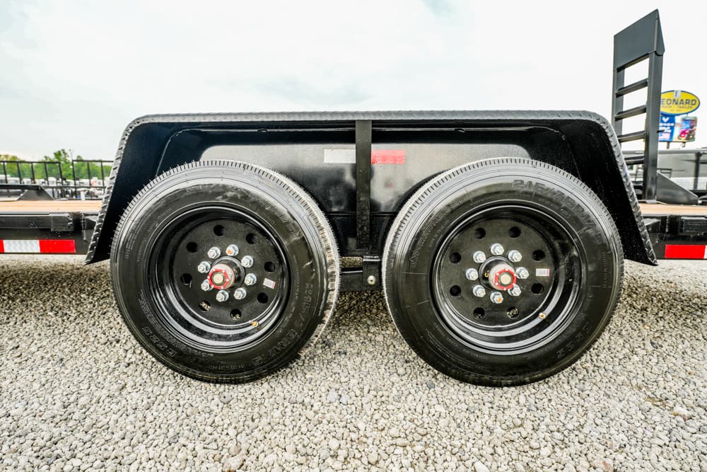 Dual-axle trailer wheels with black rims and red hub centers under a black fender on a gravel surface, close-up view. (informative)