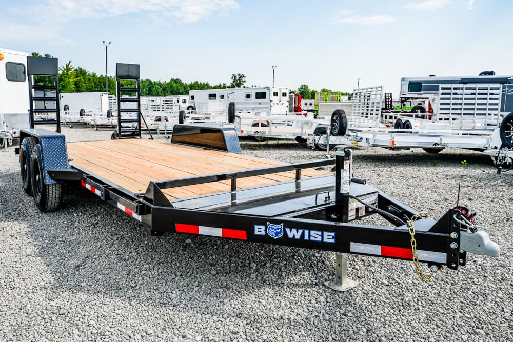 Black BowWise flatbed trailer with a wooden deck parked on a gravel lot among other trailers.