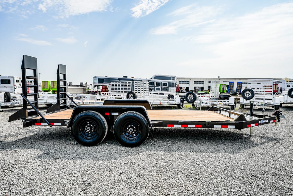 Flatbed utility trailer with dual-axle black frame and wooden deck in a gravel lot, surrounded by other trailers and equipment ramps at the rear area.