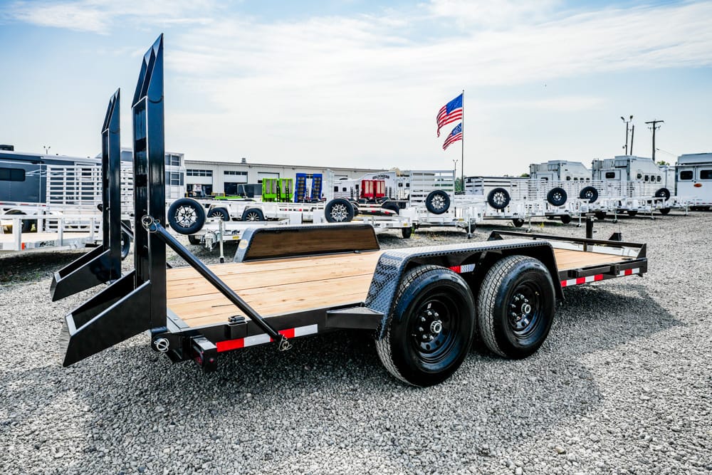 Flatbed trailer with dual axles and rear ramps on a gravel lot, surrounded by other trailers and an American flag in the background.