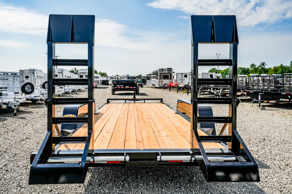 Flatbed trailer with two fold-up ramps on a gravel lot, wooden deck, surrounded by other trailers in a dealership yard