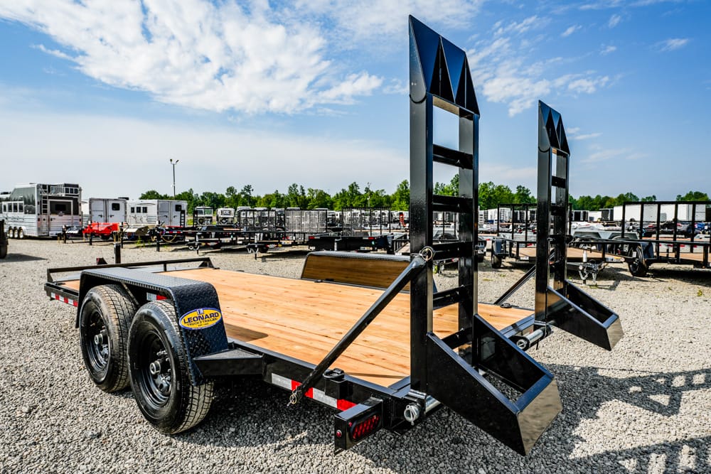 Black flatbed trailer with wooden deck and dual folding loading ramps, parked in a gravel lot among other trailers under a blue sky with scattered clouds.