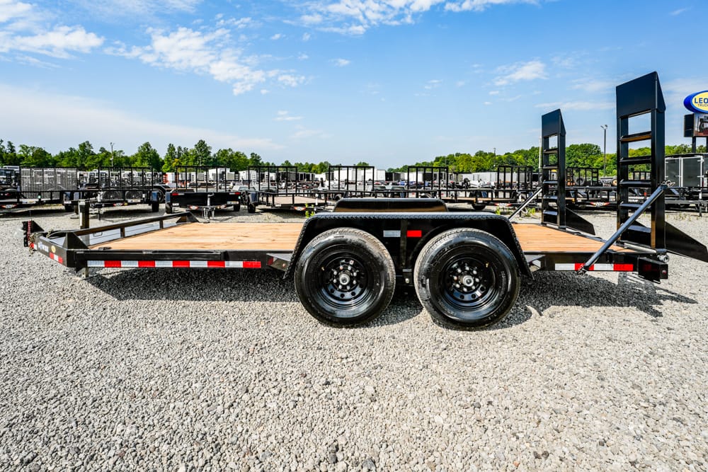 Black flatbed trailer with dual rear wheels on a gravel lot; other trailers line the background under a blue sky.