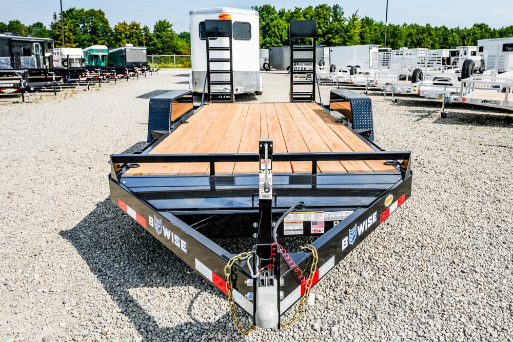 Front view of a black B-Wise flatbed trailer with a wooden deck, hitch and safety chains, parked on a gravel lot with other trailers in the background.