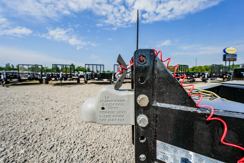 Close-up of a trailer hitch with a gray lock and red coiled safety cord on a black frame, gravel lot and trailers in the background.