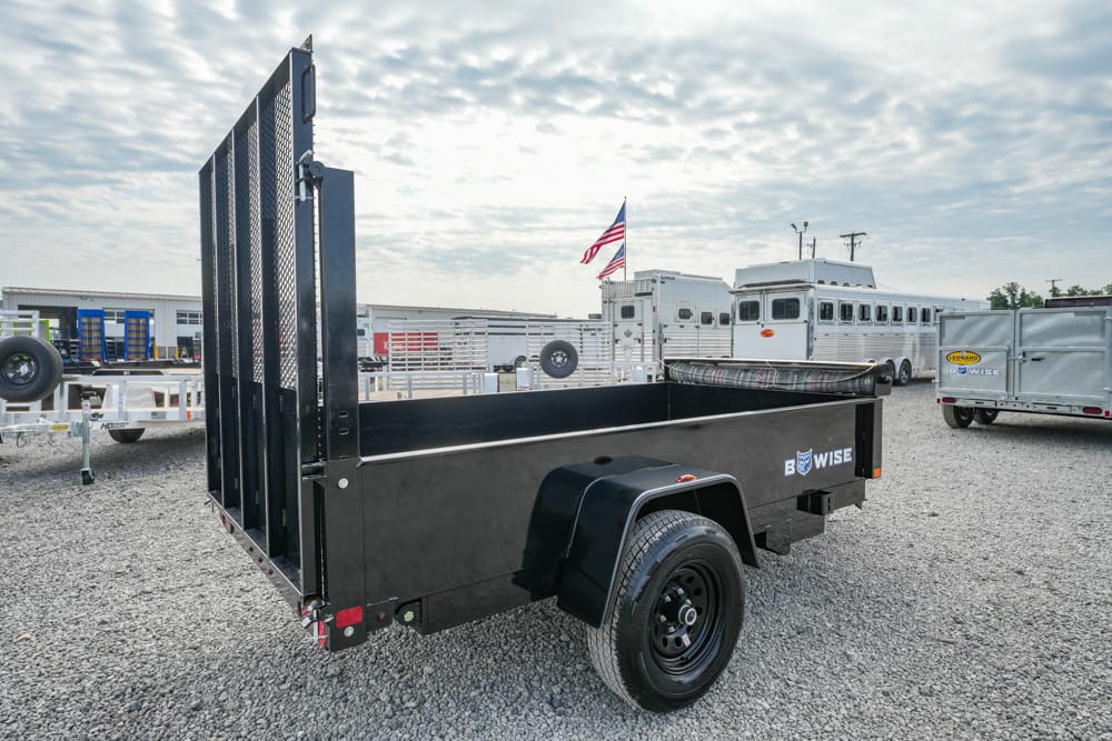 Black utility trailer with a tall ramp gate in a gravel lot, American flag visible in the background.