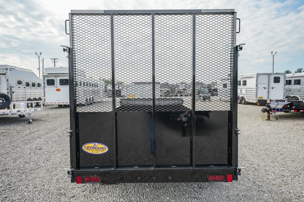 Rear view of a black utility trailer with a diamond-mesh gate, Leonard brand sticker visible, parked on a gravel lot with other trailers in the background.