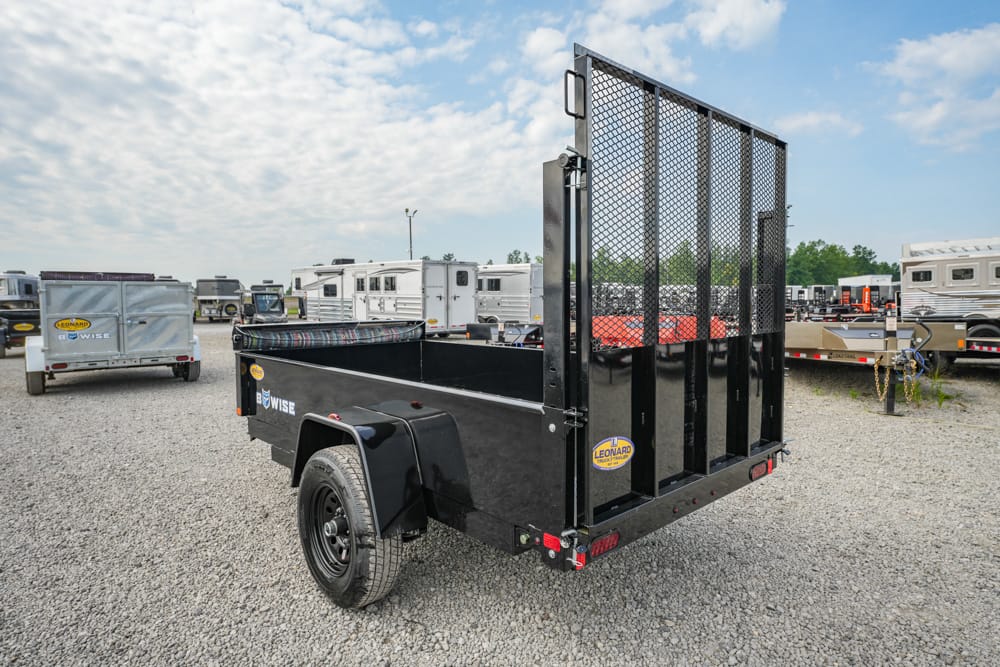Black utility trailer with a tall metal mesh gate, parked on a gravel lot among other trailers; designed for hauling equipment or materials.