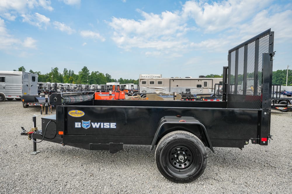 Black BowWise utility trailer with a mesh rear gate parked on a gravel lot, RVs and trailers in the background.