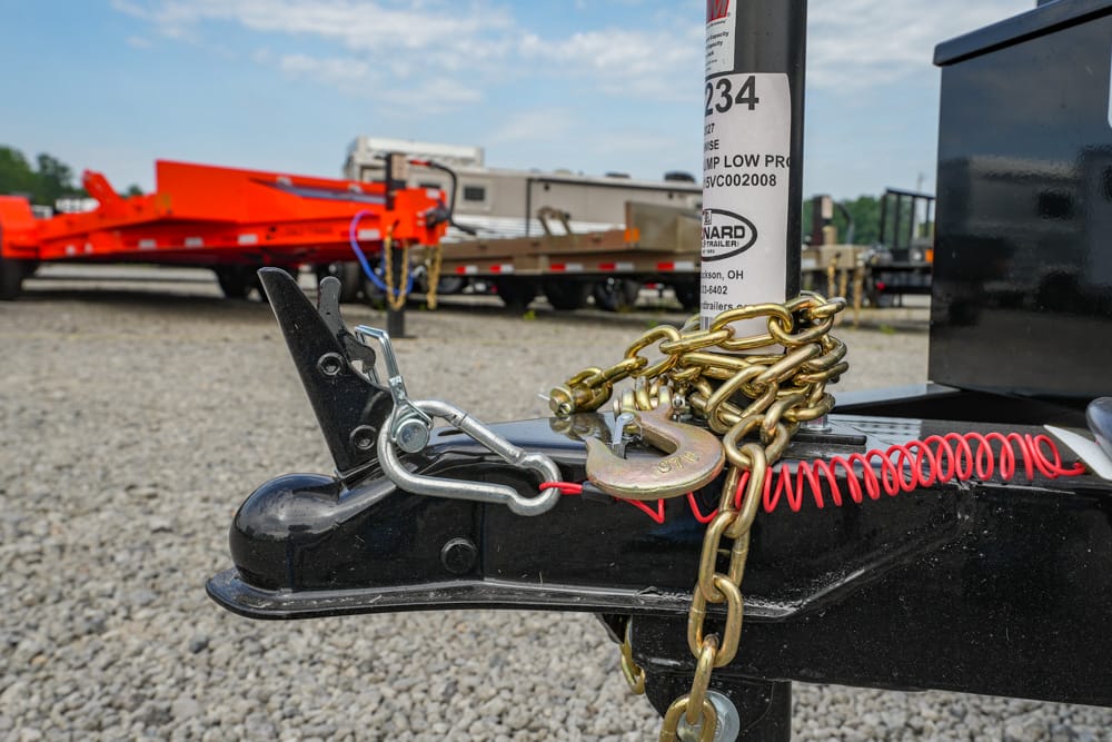 Close-up of a trailer hitch with a gold chain, padlock, and red safety wire securing a trailer on a gravel lot.