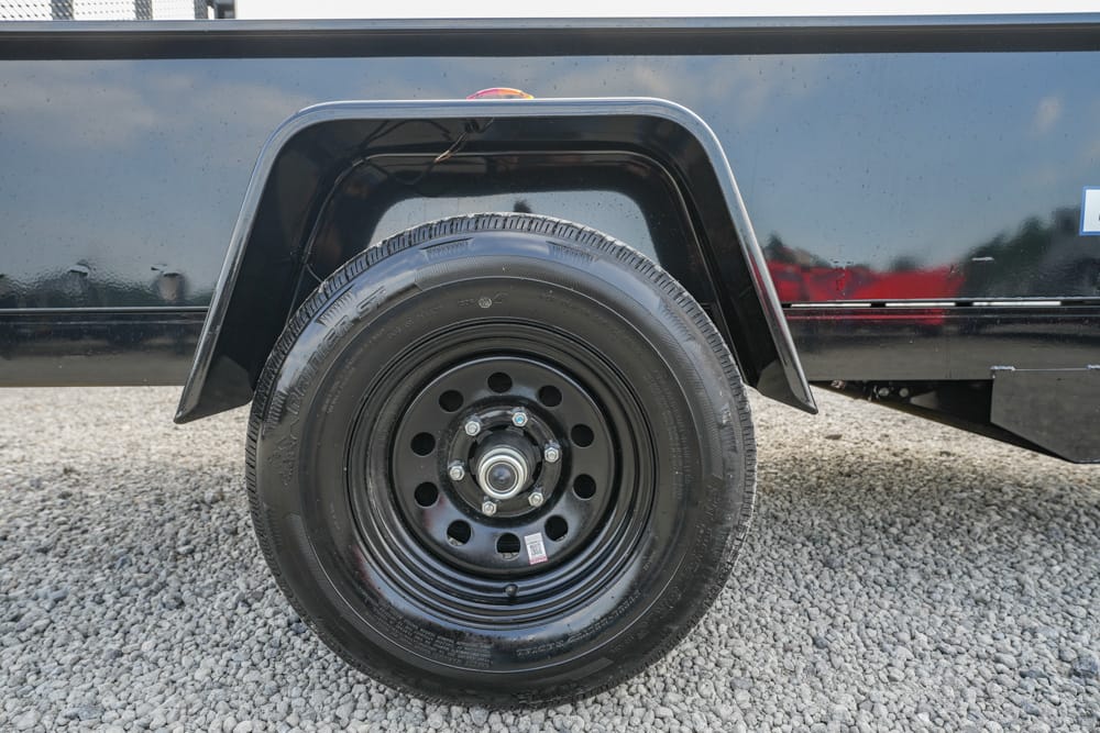 Trailer side with a black wheel and spare tire mounted in the wheel well on a gravel surface Flares and rim bolts visible, indicating a trailer tire setup.
