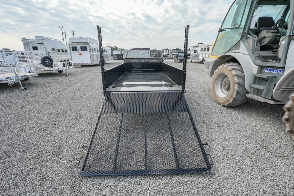 Black metal loading ramp extended from a trailer in a gravel lot, with horse trailers and a large utility vehicle nearby, ready for loading/unloading.