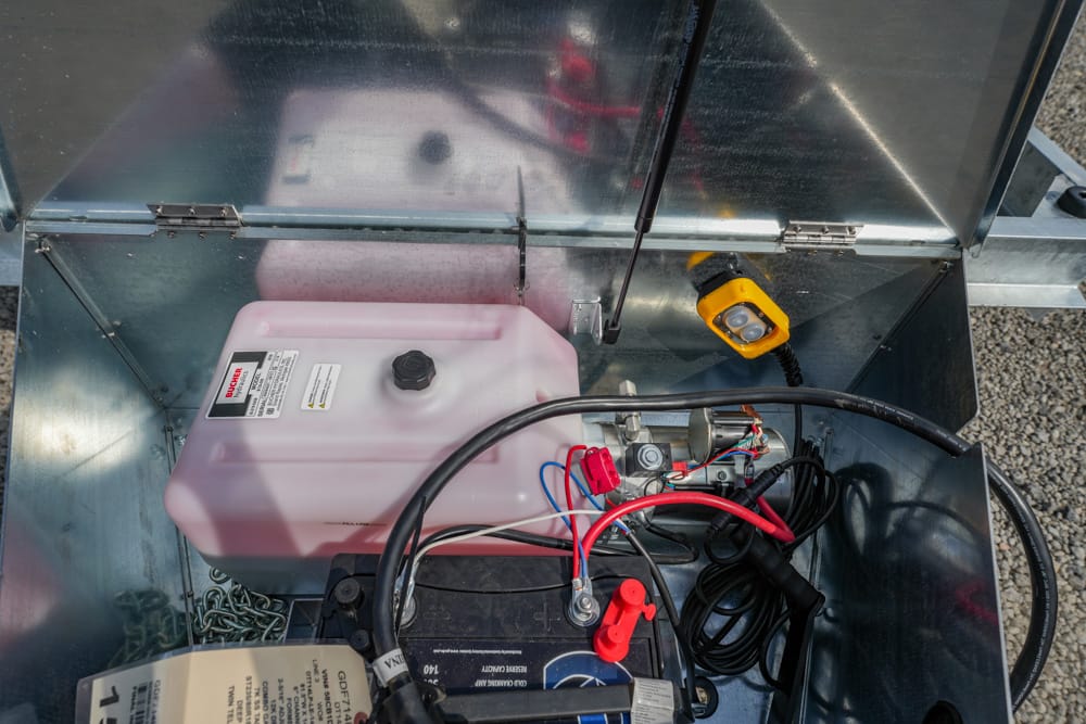Open metal compartment showing a pink translucent reservoir next to a car battery with red/black terminals and tangled cables, plus a yellow handheld work light nearby.