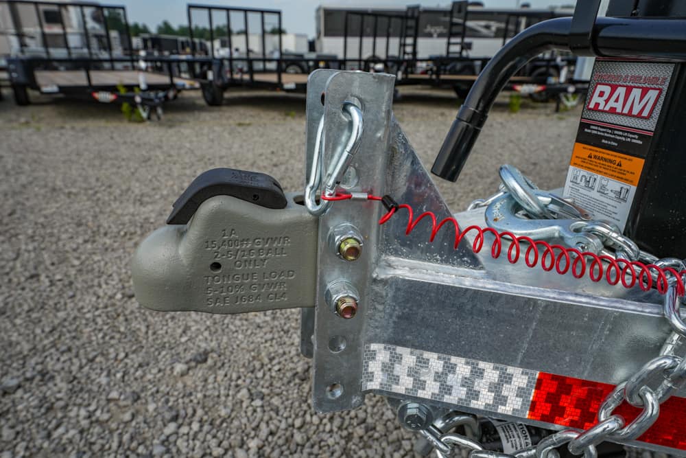 Close-up of a trailer hitch: a gray ball coupler attached to a silver frame with safety chains and a red coiled safety cable nearby, RAM labeling visible in background.