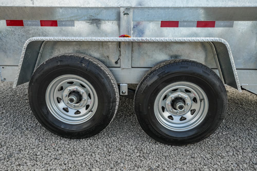Dual trailer wheels with metal fenders on a galvanized trailer resting on gravel.