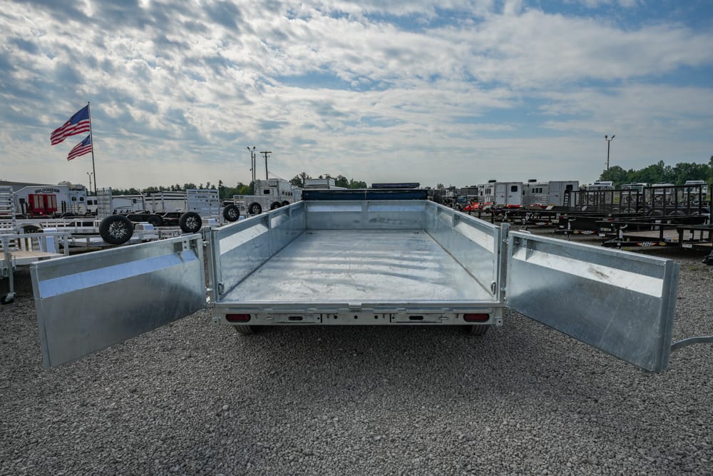 Empty aluminum utility trailer with rear ramps lowered, parked on a gravel lot under a partly cloudy sky, American flag in the background.