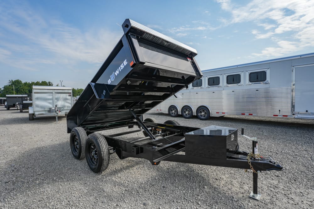 Two-axle black dump trailer with its bed raised for dumping, in a gravel lot beside other cargo trailers