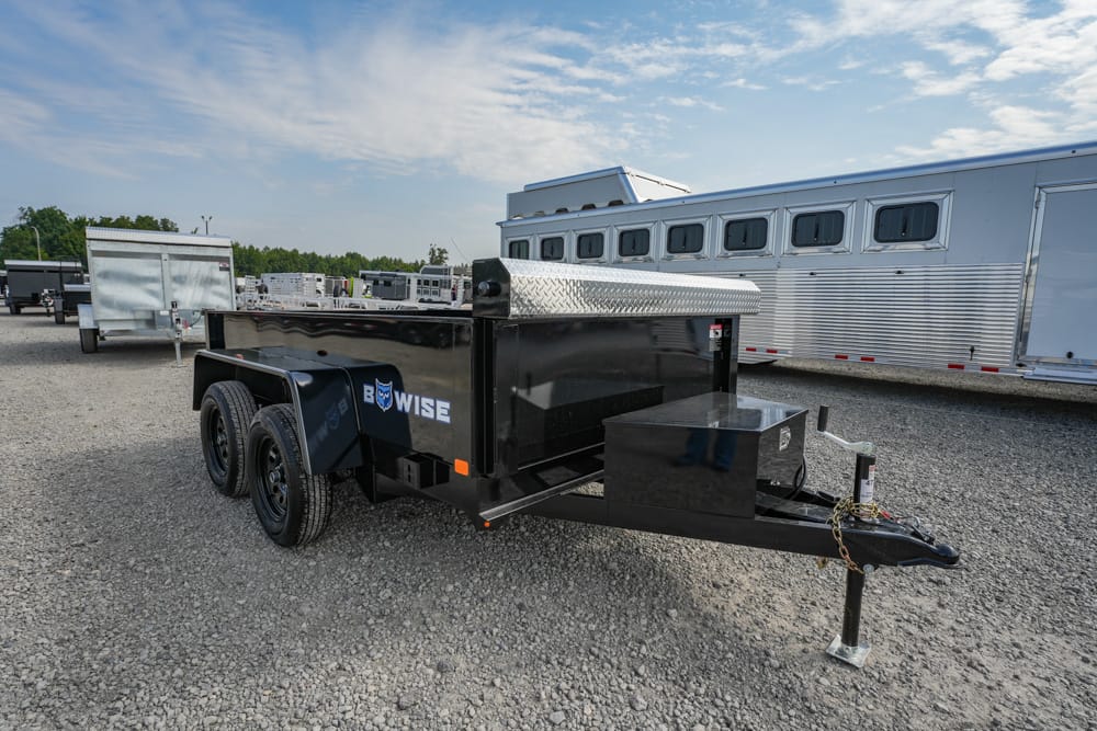 Black dual‑axle dump trailer with diamond-plate toolbox, parked on a gravel lot with silver horse trailers in the background.