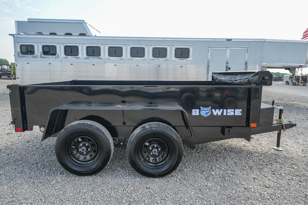 Black dual-axle utility trailer with a coiled hose reel on top, parked on gravel, logo 'B WISE' on the side, and a silver horse trailer in the background.