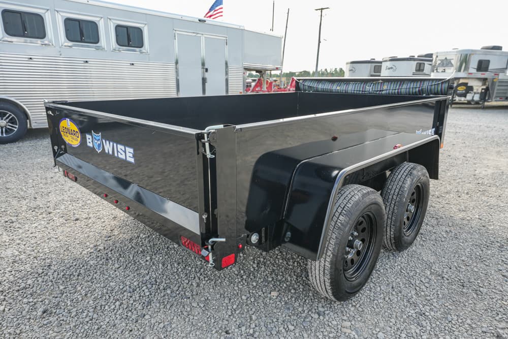 Black dual-axle dump trailer on a gravel lot with other trailers in the background.