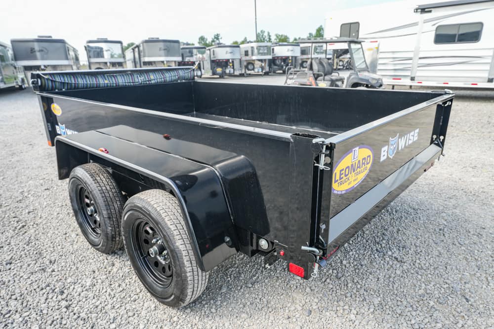 Black double-axle dump trailer on a gravel lot with RVs in the background.