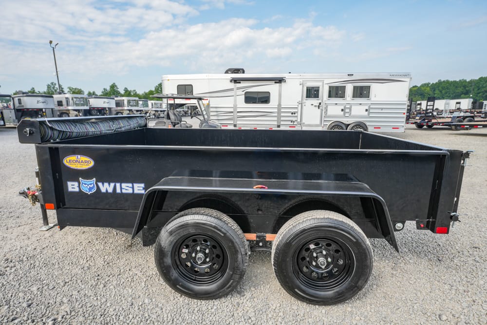 Black Bowwise dump trailer with dual axles in a gravel lot, surrounded by other horse trailers in the background