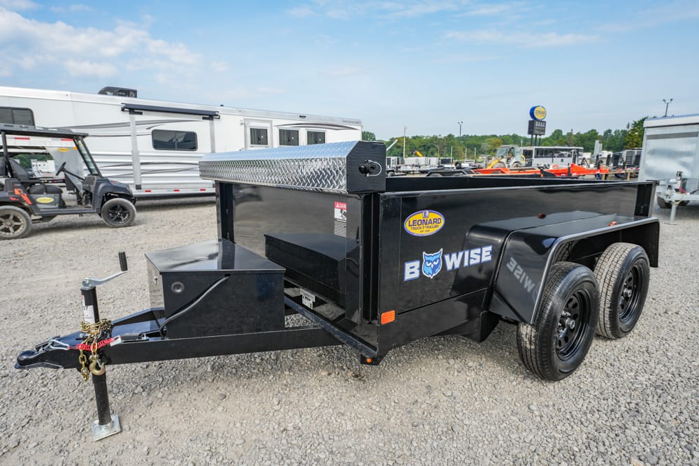 Black dual-axle BowWise utility trailer on a gravel lot, with a diamond-plate toolbox and hitch in the foreground.