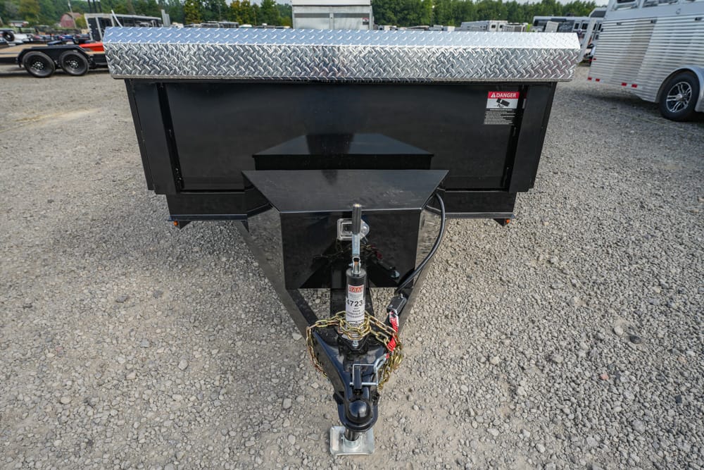 Black dump trailer hitch and front storage box on a gravel lot with diamond-plate trim top edge.